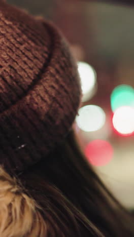 vista en primera persona de una mujer con un abrigo de invierno con una capucha de piel, dentro de un coche por la noche. ella está ajustando su calentador de cabeza en su cabeza con los dedos pintados de negro. el fondo muestra luces borrosas de la ciudad