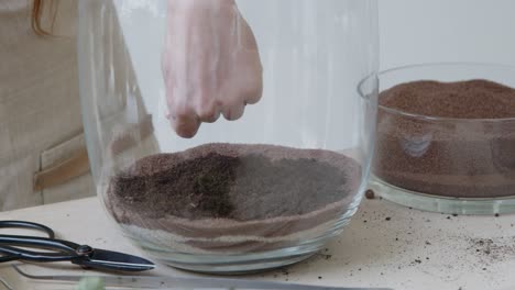 A-young-female-botanist-creates-a-tiny-live-forest-ecosystem-in-a-huge-glass-jar---the-main-soil-layer-putting-by-hand---a-tight-close-up