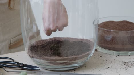 a young female botanist creates a tiny live forest ecosystem in a huge glass jar - the main soil layer putting by hand - a tight close-up