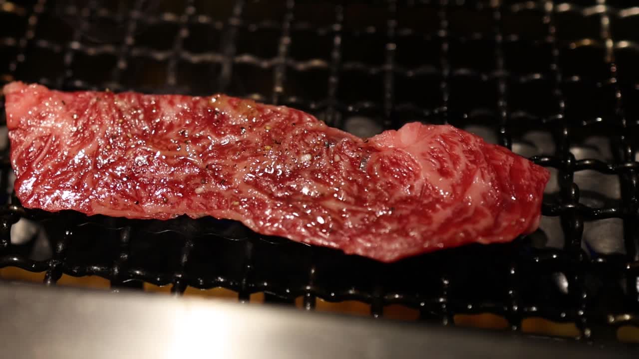 Close-up of marbled beef strips being placed and grilled over an open flame on a metal grate.