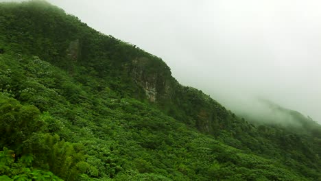 Mist-coming-over-the-top-of-a-cliff,-which-is-part-of-a-mountain-range-covered-in-vegetation