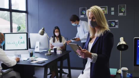 Diverse-business-colleagues-wearing-face-masks-using-digital-tablet-and-talking-in-office