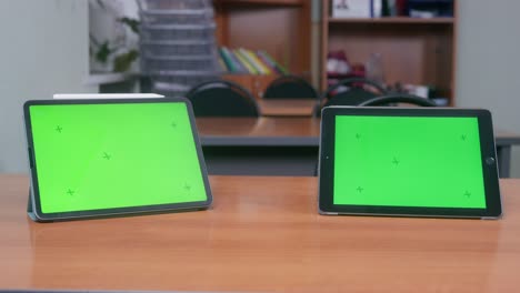 pair of black tablets with chromakey screens stands on wooden table against shelves with school supplies and grey wall in classroom