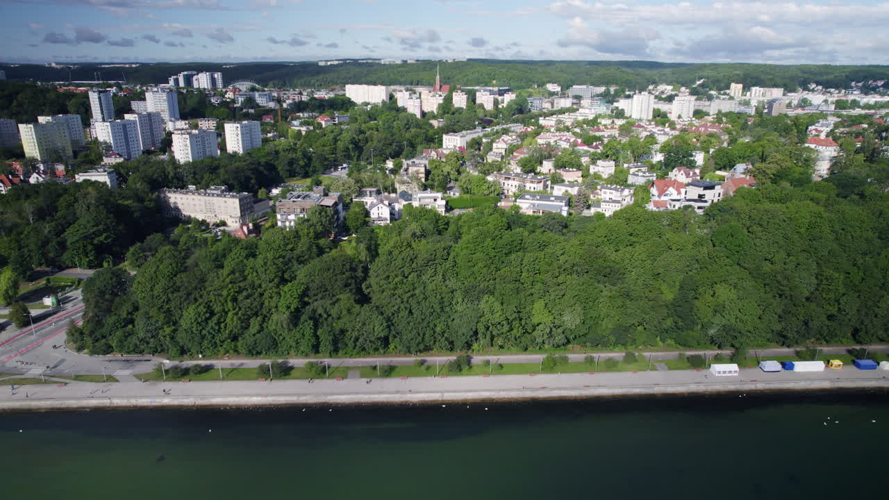vista aérea, volando a lo largo del bulevar costero gdynia polonia, agua de mar verde, bulevar de la calle a lo largo de la costa, colina cubierta de bosque y paisaje urbano de gdynia en el fondo