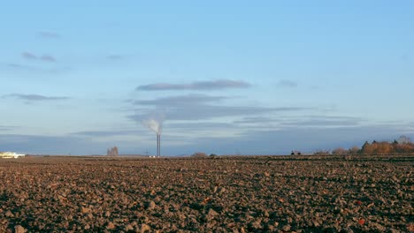 An-empty-brown-field-with-people-walking-on-the-horizon-and-a-working-chimney