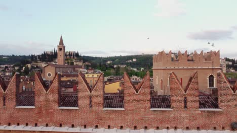 drone pans right to left over verona red brick battlements, revealing the tall bell tower of sant’anastasia church and cypress-covered castel san pietro hill, with renaissance villas in the background