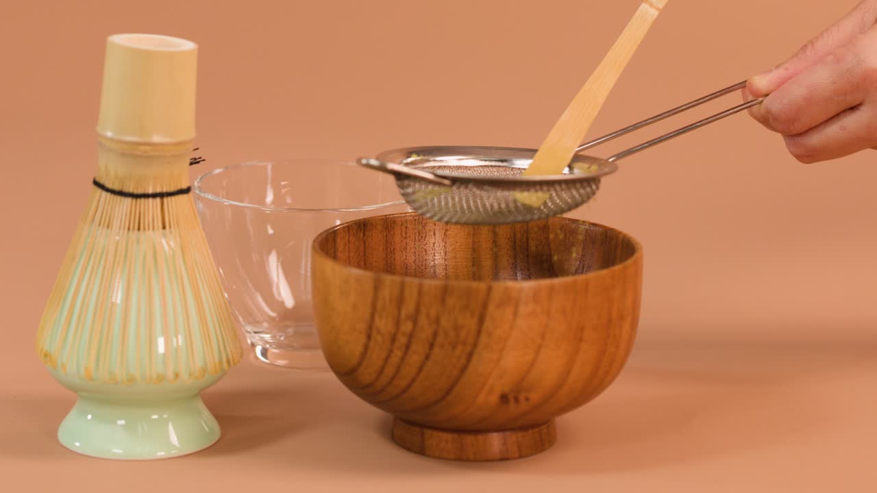Hand sifts matcha powder through strainer into wooden bowl, soft lighting, neutral background, static camera