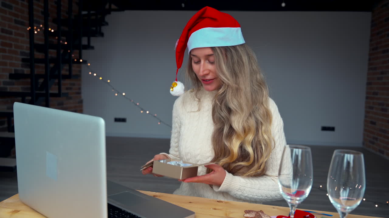 A woman in a Christmas hat opens a gift while sitting at a rustic table. Soft lights create a warm atmosphere as she smiles and enjoys her festive time indoors
