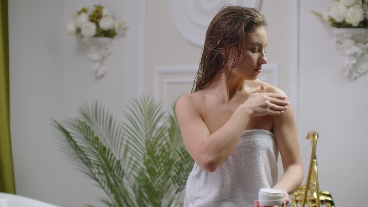 hermosa mujer joven en toalla de baño aplicando loción corporal en sus hombros y sonriendo mientras está de pie en el baño