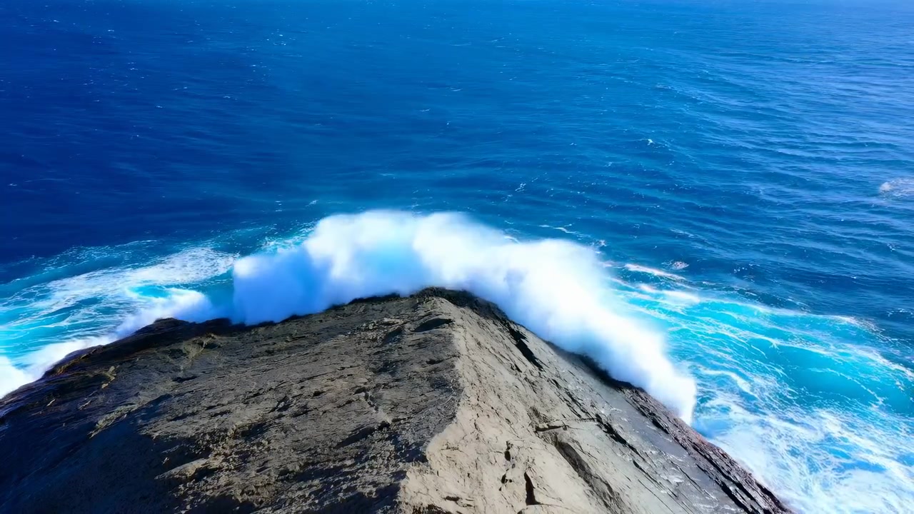 Elevated view follows rolling swell as it builds behind exposed rock, breaks over the ridge, and sends dense white spray and turquoise foam across rugged stone under clear daylight.