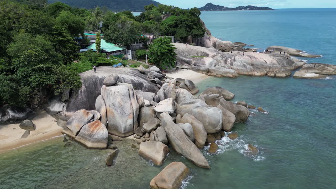 Left to tight pan shot of a pile of huge rocks placed at the beach with clear clean water and lush green trees in the background