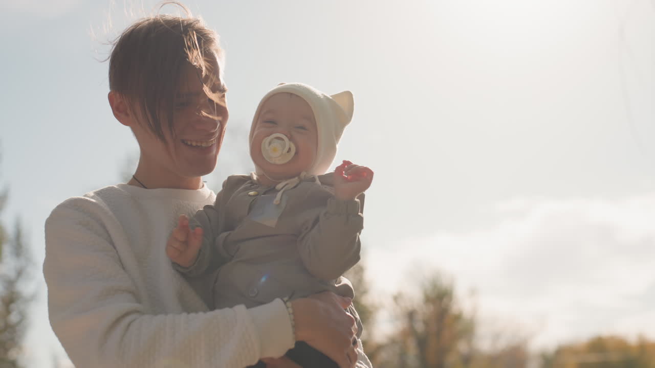 Hvid far holder baby udendørs om efteråret. Blødt modlys nær et grønt hegn, strikhue med ører, sut, blidt smil, tæt omfavnelse, ægte omsorgsøjeblik, forældrevarme og familie.
