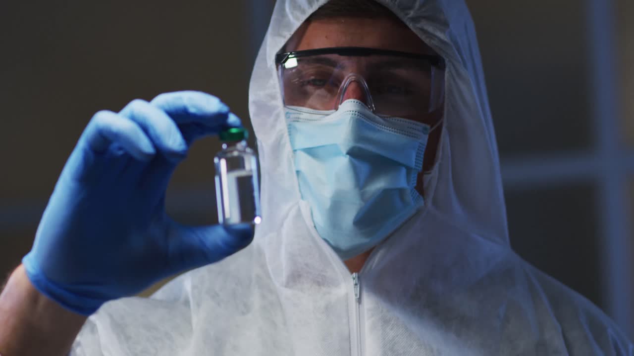Trabajador médico caucásico con ropa protectora, mascarilla y guantes sosteniendo un vial de vacuna en el laboratorio