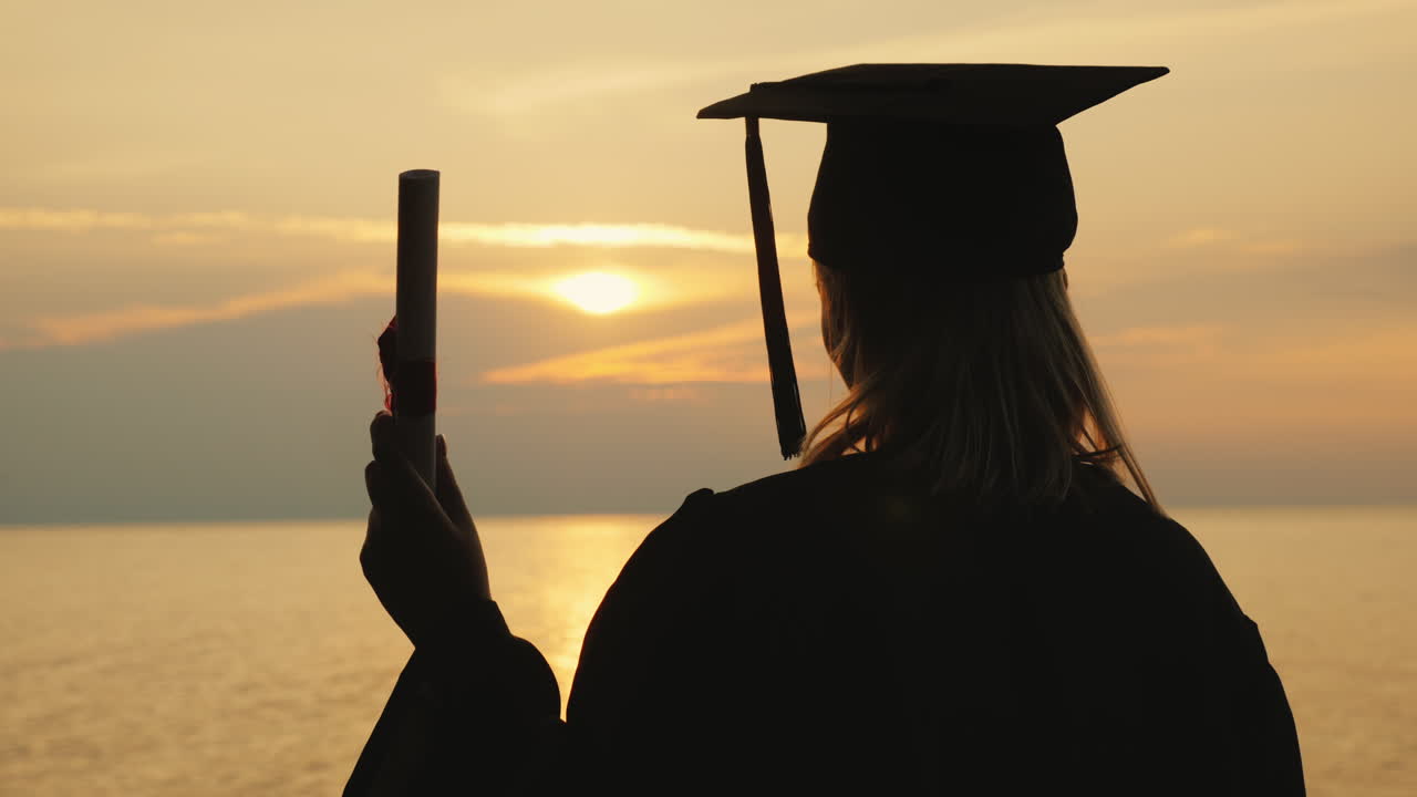 un soltero con un diploma en la mano y una gorra de graduado mira el amanecer sobre el mar