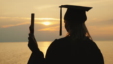 a bachelor with a diploma in hand and a cap of a graduate looks at the sunrise over the sea