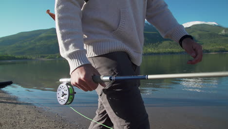 close up tracking shot of a fly fisherman's rod and reel while walking in front of gorgeous, snow-capped mountains, and a deep, reflective mountain lake