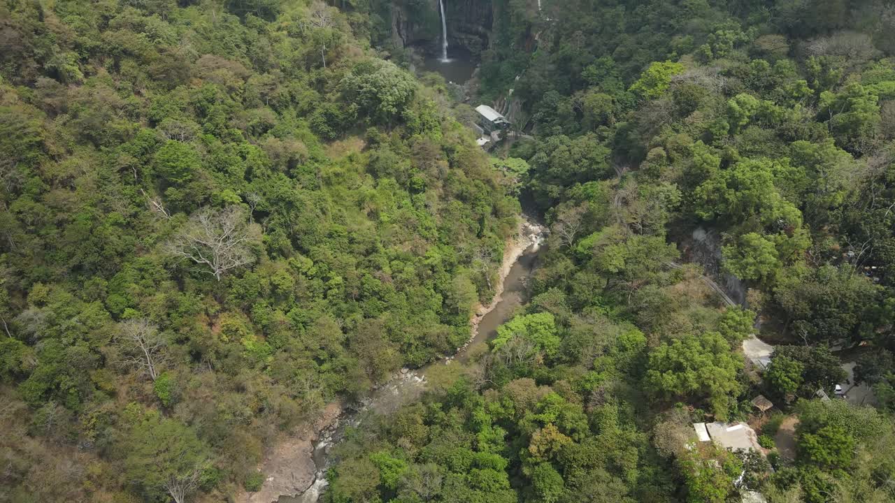 Scenic aerial view of Indonesian jungle landscape with a hidden waterfall dropping from a rocky cliff into a natural basin, framed by dense rainforest and green tropical valley