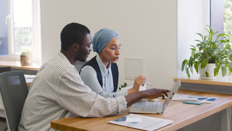 Young-Worker-Working-With-Laptop-Sitting-At-His-Desk-While-Muslim-Businesswoman-Talks-To-Him-Sitting-At-Desk-While-Taking-Notes