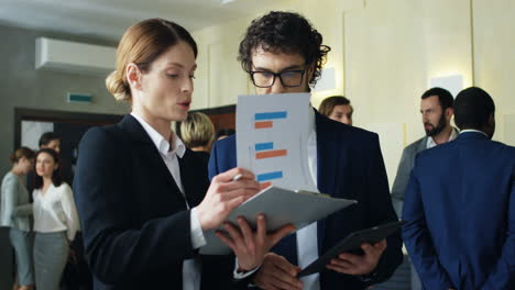 caucasian businesswoman reading documents and charts and her male colleague holding a tablet while they talking and discussing something in a conference hall