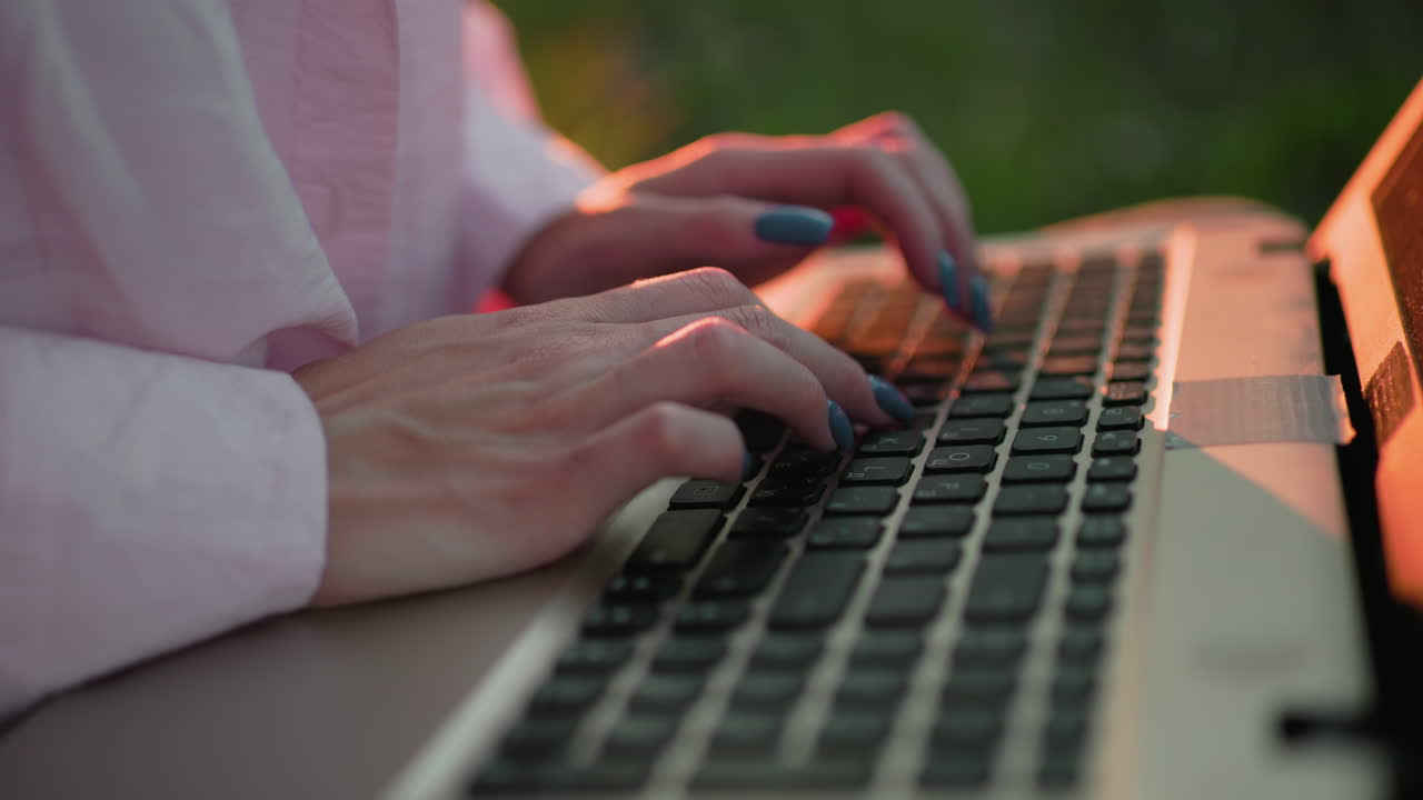 primer plano de una mujer con una camiseta rosa con uñas bien pulidas escribiendo en una computadora portátil, sombras de su mano visibles en el teclado, con un cálido resplandor de luz solar en el fondo y un efecto de luz bokeh en la distancia