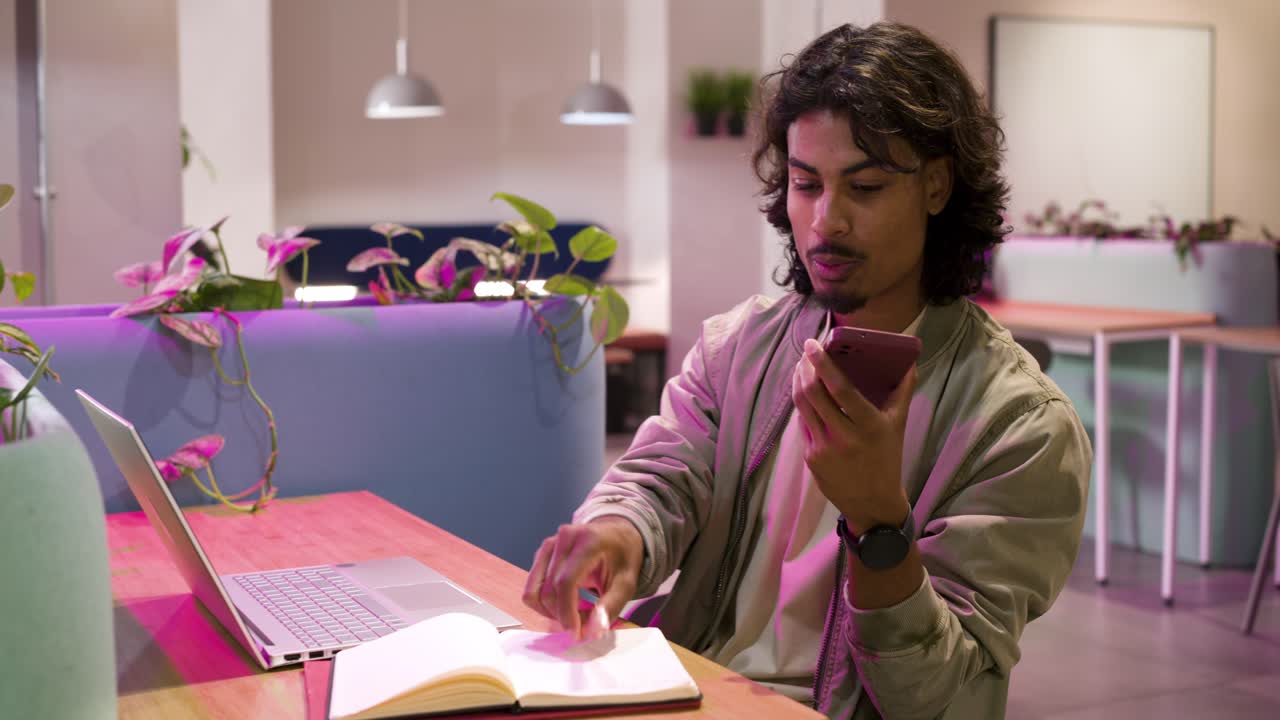 Indian man in jacket receiving phone alert checking it and noting notebook for work at workspace