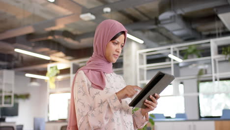 happy biracial casual businesswoman in hijab using tablet in office, slow motion