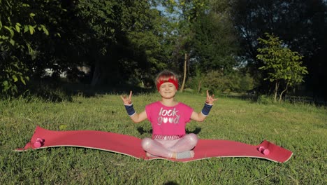 child sitting on mat and performing yoga meditation outdoors in park. girl doing yoga exercises