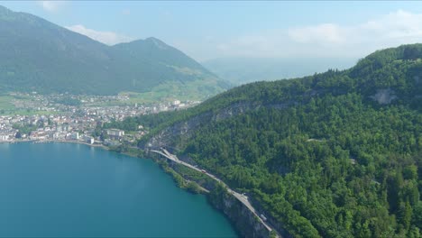 Paso-Elevado-De-Morschach:-Moviéndose-Sobre-El-Pintoresco-Valle-De-Agua-Hacia-Las-Montañas-De-Los-Alpes,-Suiza,-Europa,-Drone-|-Movimiento-Más-Cercano-Hacia-El-Interior-A-Lo-Largo-De-La-Carretera-Junto-Al-Acantilado-Junto-Al-Majestuoso-Lago-Azul