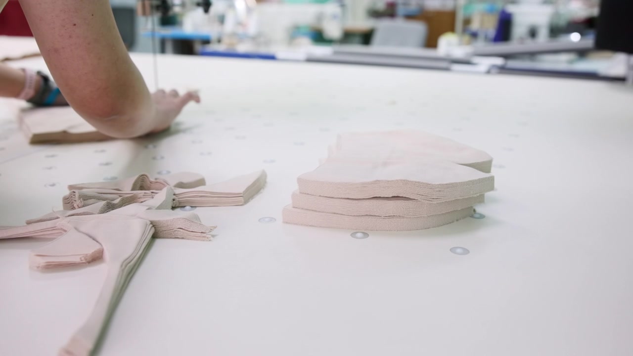 Hands Pushing Stack Of Cut Fabric Pieces Across Large Cutting Table, Busy Workshop Backdrop With Tools . Media
