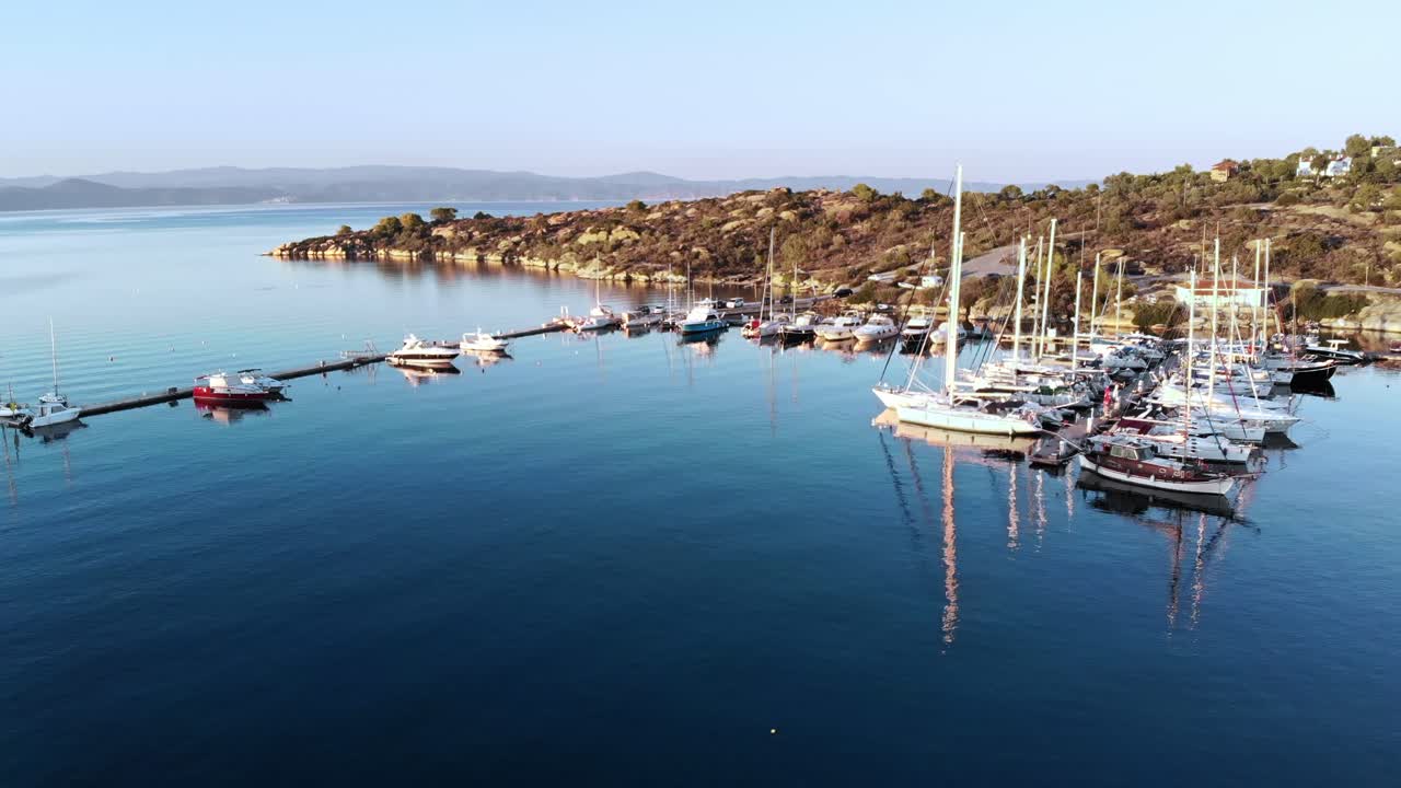 Aerial drone view of Aegean sea port with multiple moored yachts at piers, greenery, blue water, sunset, Greece