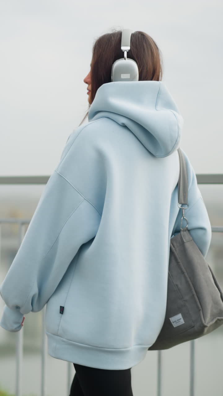 Side view of beautiful girl in casual hoodie walking with bag near iron rail, blurred view of bridge and river in background, creating a calm, urban atmosphere perfect for lifestyle content