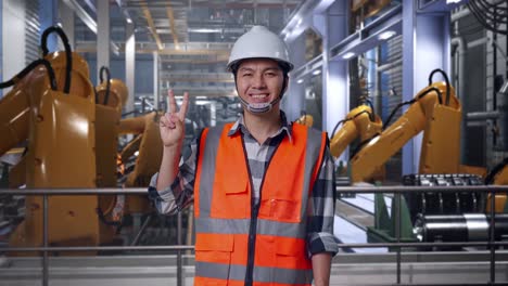 asian male engineer with safety helmet smiling and showing peace gesture while standing with industrial robotic arms working