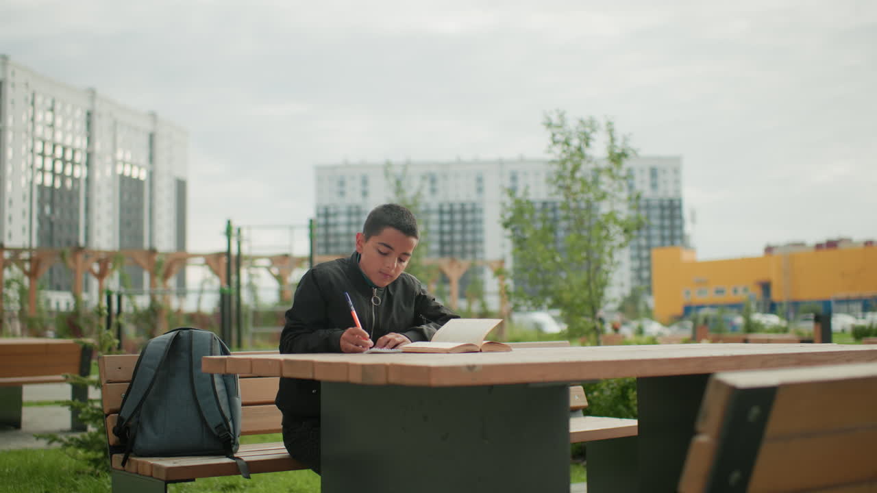 Teen boy in black jacket writes in notebook with pen beside open book on wooden table, studying with focus outdoors in urban setting, slight blur background of buildings and trees