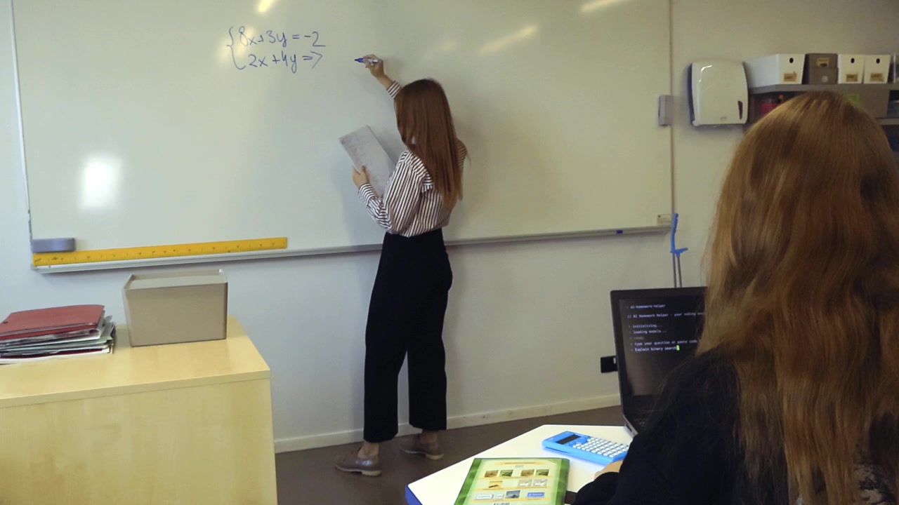 Insegnante di matematica donna che scrive un sistema di equazioni lineari su una lavagna bianca mentre uno studente utilizza un programma di intelligenza artificiale su un computer portatile per trovare la soluzione