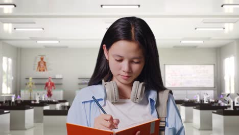 close up of asian teen girl student with a backpack smiling and taking note on notebook while standing in science laboratory