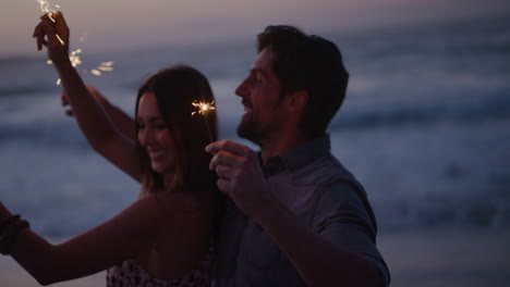 joven pareja feliz agitando chispas bailando celebrando la víspera de año nuevo en la playa tranquila puesta de sol de fondo chispas de fuegos artificiales