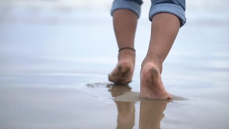 siguiendo los pies de un bebé indefinido caminando en la playa con sus padres en la tarde con olas de mar salpicando sus piernas descalzas, cámara lenta
