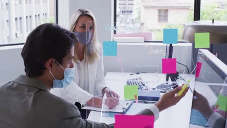 Diverse-business-colleagues-wearing-face-masks-using-computers-talking-in-office