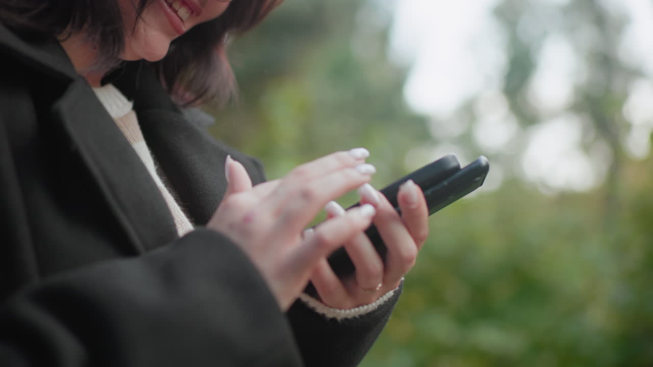 자연을 좋아하는 사람이 두 개의 휴대폰을 들고 매니큐어를 칠한 손톱으로 맨 위에 있는 화면을 터치하는 모습이에요. 밝은 표정을 짓고 있고, 공원에서 아늑한 코트를 입고 있으며, 부드러운 보케 배경에 현대적인 소통을 하고 있어요.