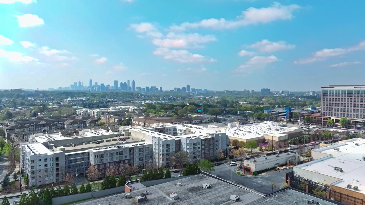 Atlanta suburban apartment complex with flat white rooftops, mixed-use buildings, tree-lined streets and distant downtown skyline under partly cloudy blue sky. Georgia, USA. Aerial wide shot.