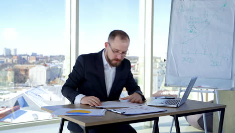 nervous businessman in office. irritated man entrepreneur sitting at table and tearing papers throwing them to the sides on the background of window.