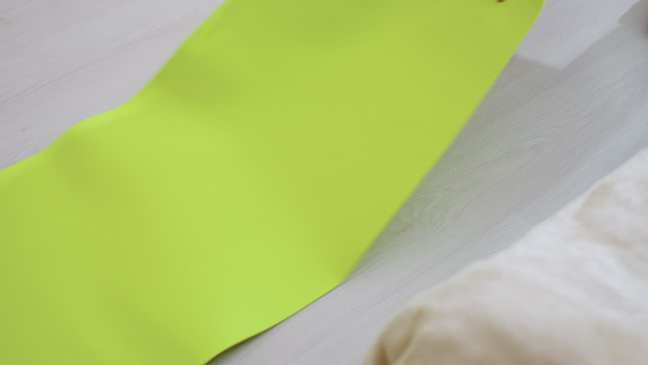 Person wearing light socks and dark pants begins step onto vibrant green fitness mat on pale wooden floor, casting distinct shadow in soft natural light, preparing for indoor workout