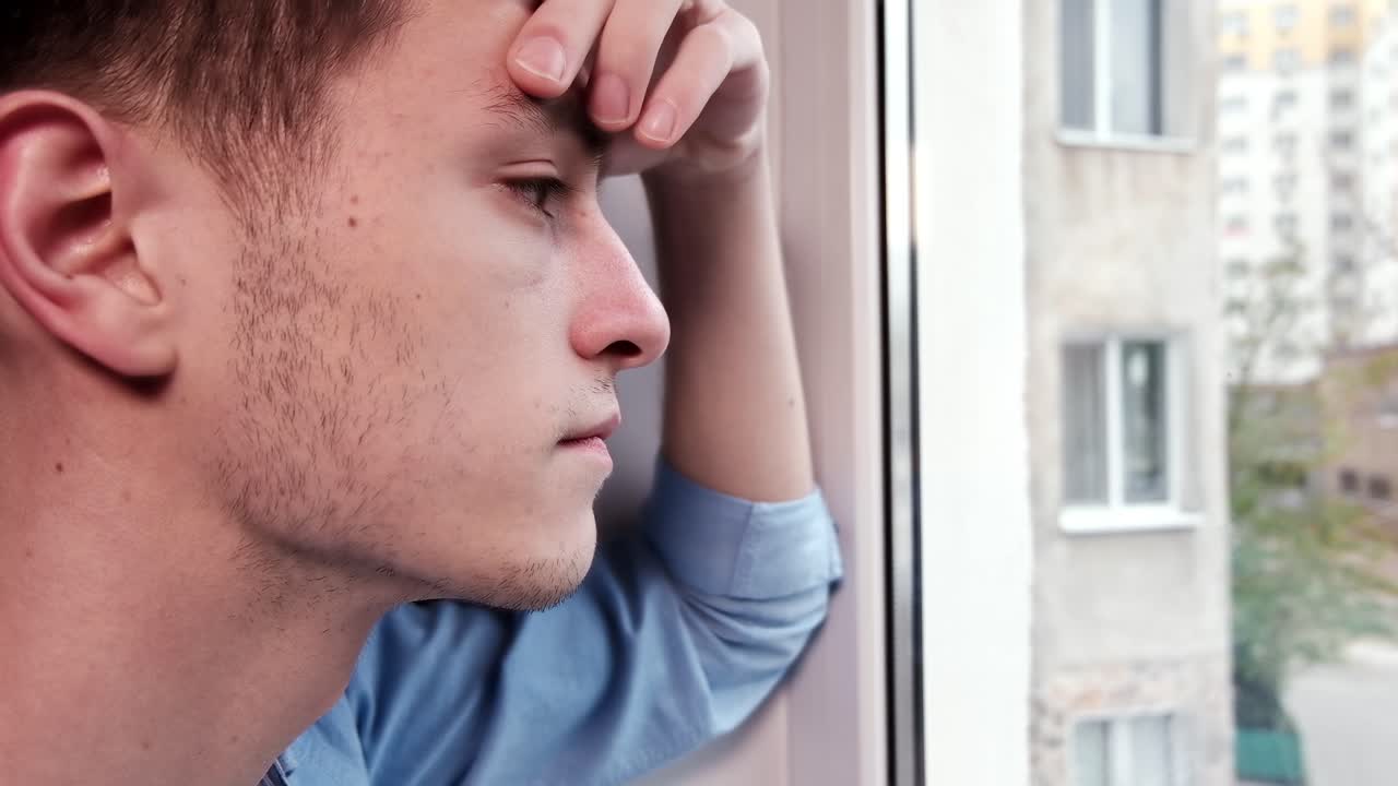 Bored man in blue shirt looking desperate out of the window. Close up. Quarantine and Corona Virus idea