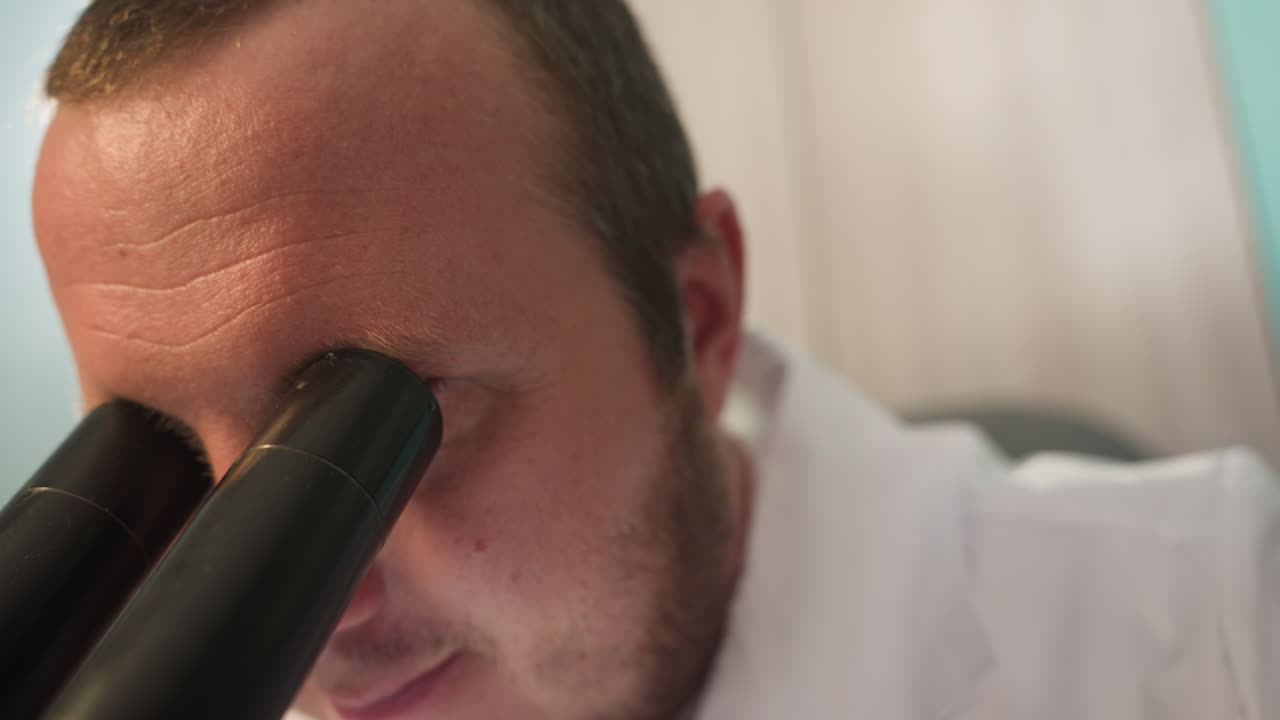 una vista de cerca de un técnico en una bata de laboratorio blanca examinando algo bajo un microscopio, la cara del técnico está ligeramente borrosa, centrándose en el microscopio en primer plano