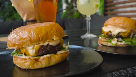 two gorgeous and tasty gourmet burgers or hamburgers on a restaurant table outdoors with some drinks in the background. burgers are in focus, background blurred. meat is juicy and sauce is dripping.