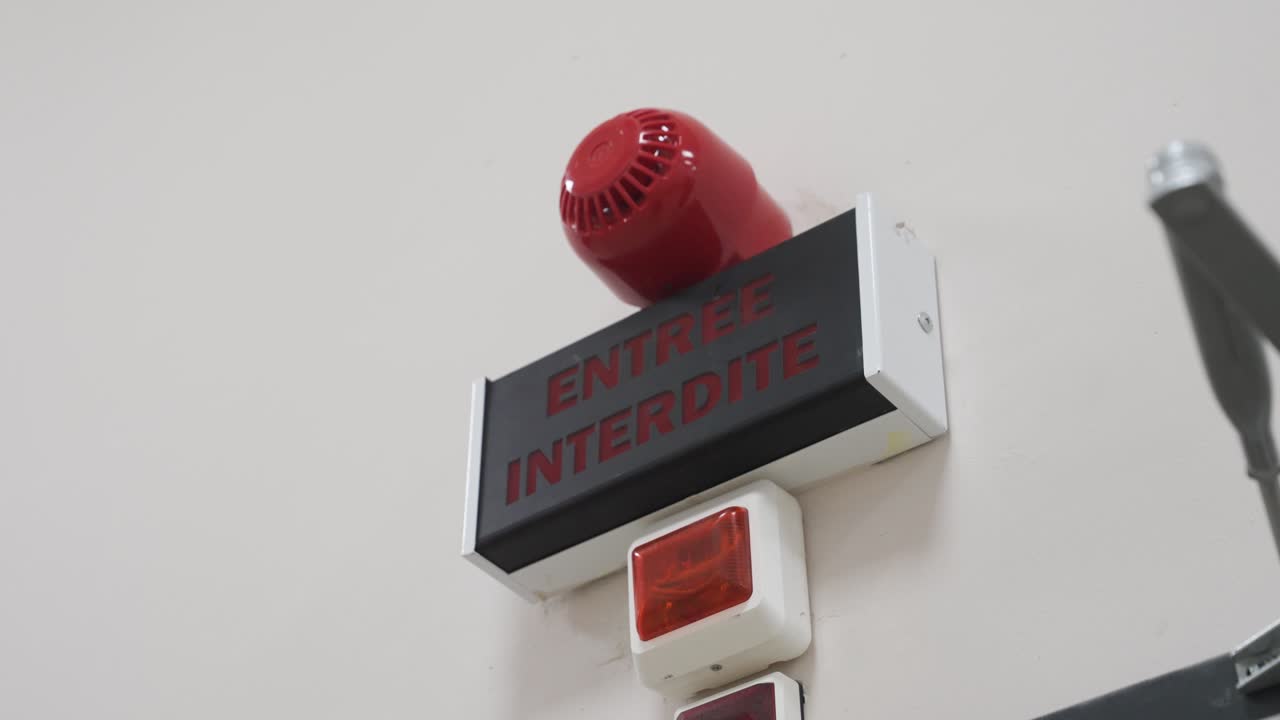 A close-up on a wall-mounted warning system for a restricted area. A sign reads "ENTREE INTERDITE" (Do Not Enter) and is fitted with an alarm siren and flashing light