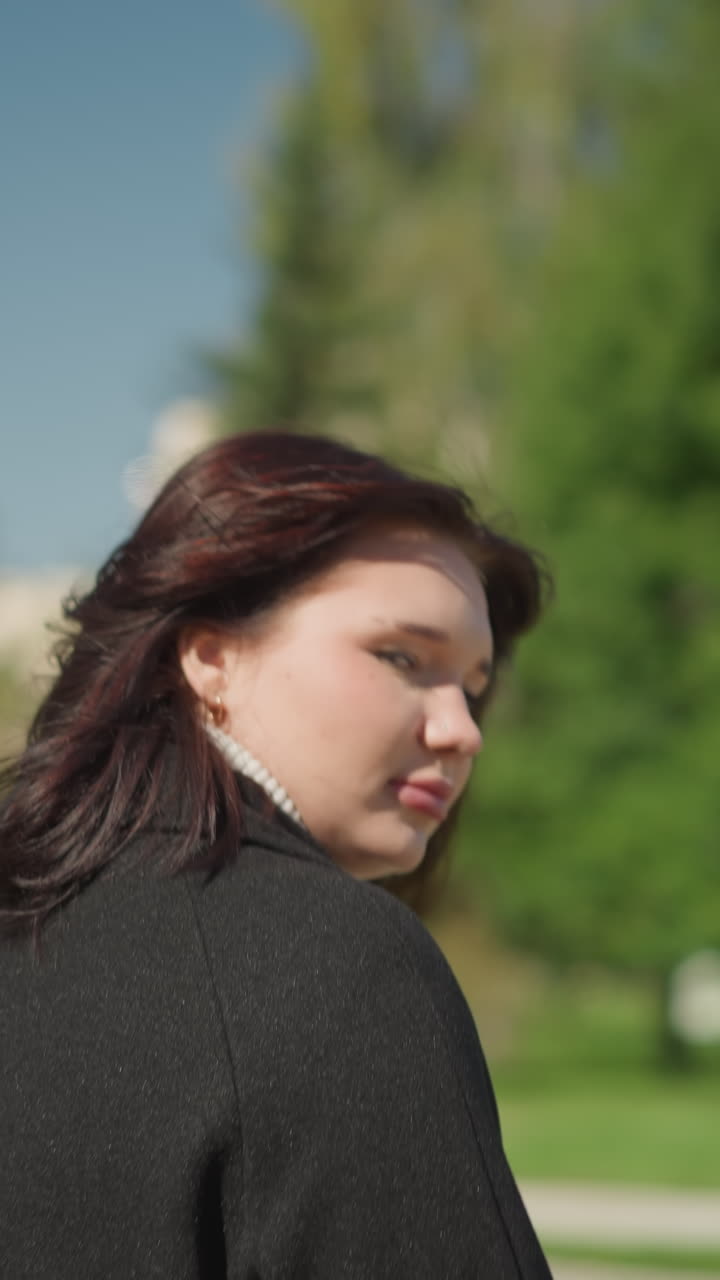 검은 옷을 입은 여성이 야외에서 빛 아래 머리카락을 조정하는 동안, 활기찬 초록색, 경로, 그리고 공공 공원에서 멀리 있는 나무와 함께