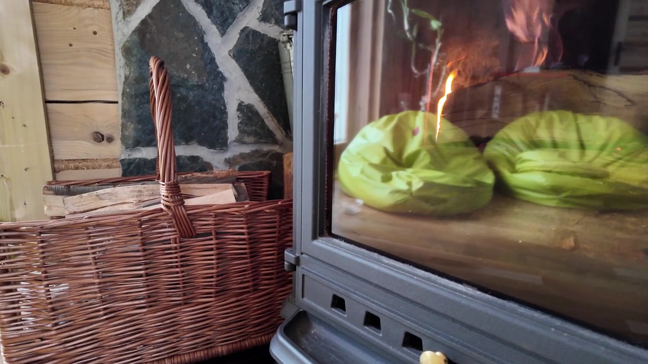 Una stufa a legna riscaldante ripresa in dettaglio, che mostra fiamme danzanti e riflessi interni, con un cesto di vimini pieno di legna da ardere.