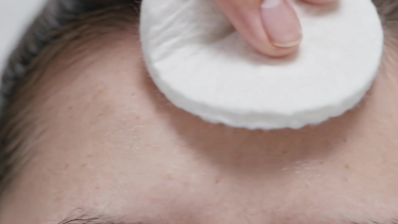 Pressing cotton pad onto hairline, hand wiping subject's forehead at vanity, removing oily residue