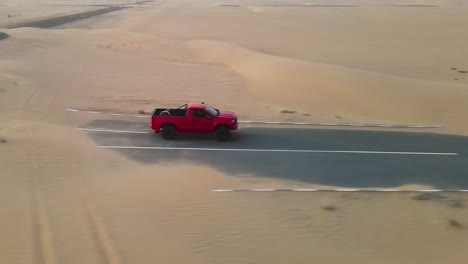 Camión-Rojo-Brillante-Que-Conduce-A-Una-Sola-Carretera-Con-Arena-Que-Se-Derrama-Desde-El-Desierto,-Rastreo-De-Drones-Desde-El-Costado